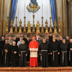 Grupa duchownych w czarnych habitach, z kardynałem w czerwieni, pozuje przed ołtarzem. Wszyscy razem śpiewają "Ciebie Boga wysławiamy".