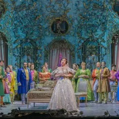 Primadonna w białej sukni śpiewa na scenie operowej, otoczona przez barwnie ubranych artystów w bogato zdobionej, błękitnej scenografii.