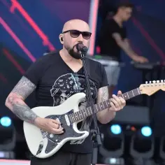 Łysy gitarzysta w okularach przeciwsłonecznych gra na białej gitarze. Na scenie, za nim, stoi klawiszowiec. To koncert, gdzie grają kombi piosenki alfabetycznie.