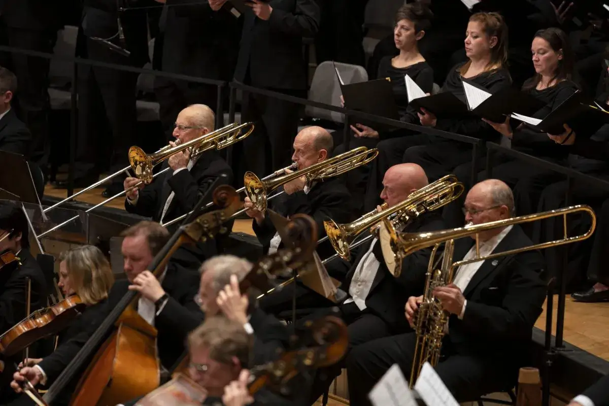 Orkiestra gra na instrumentach dętych blaszanych. Widać puzony i inne instrumenty, a także muzyków w czarnych strojach.