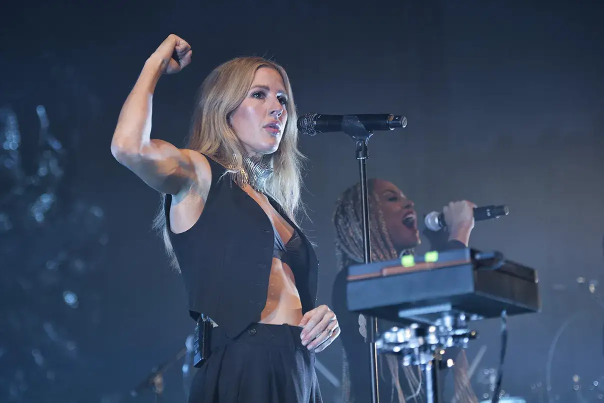 Ellie Goulding na koncercie, z podniesioną pięścią, śpiewa swoje utwory. W tle wokalistka z warkoczami.