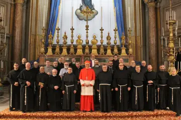 Grupa duchownych w czarnych habitach, z kardynałem w czerwieni, pozuje przed ołtarzem. Wszyscy razem śpiewają "Ciebie Boga wysławiamy".