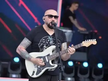 Łysy gitarzysta w okularach przeciwsłonecznych gra na białej gitarze. Na scenie, za nim, stoi klawiszowiec. To koncert, gdzie grają kombi piosenki alfabetycznie.