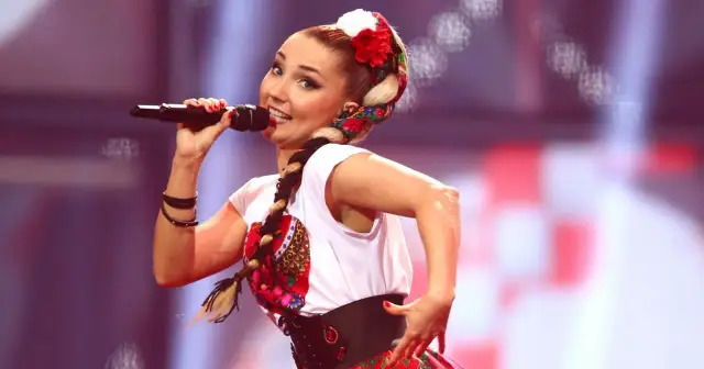 Cleo na Eurowizji w ludowym stroju, z warkoczem i kwiatami we włosach, śpiewa do mikrofonu.