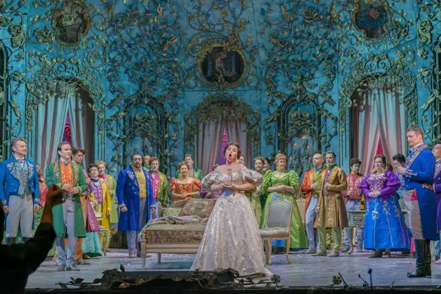 Primadonna w białej sukni śpiewa na scenie operowej, otoczona przez barwnie ubranych artystów w bogato zdobionej, błękitnej scenografii.