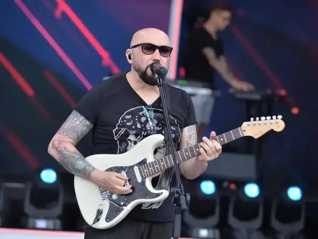 Łysy gitarzysta w okularach przeciwsłonecznych gra na białej gitarze. Na scenie, za nim, stoi klawiszowiec. To koncert, gdzie grają kombi piosenki alfabetycznie.